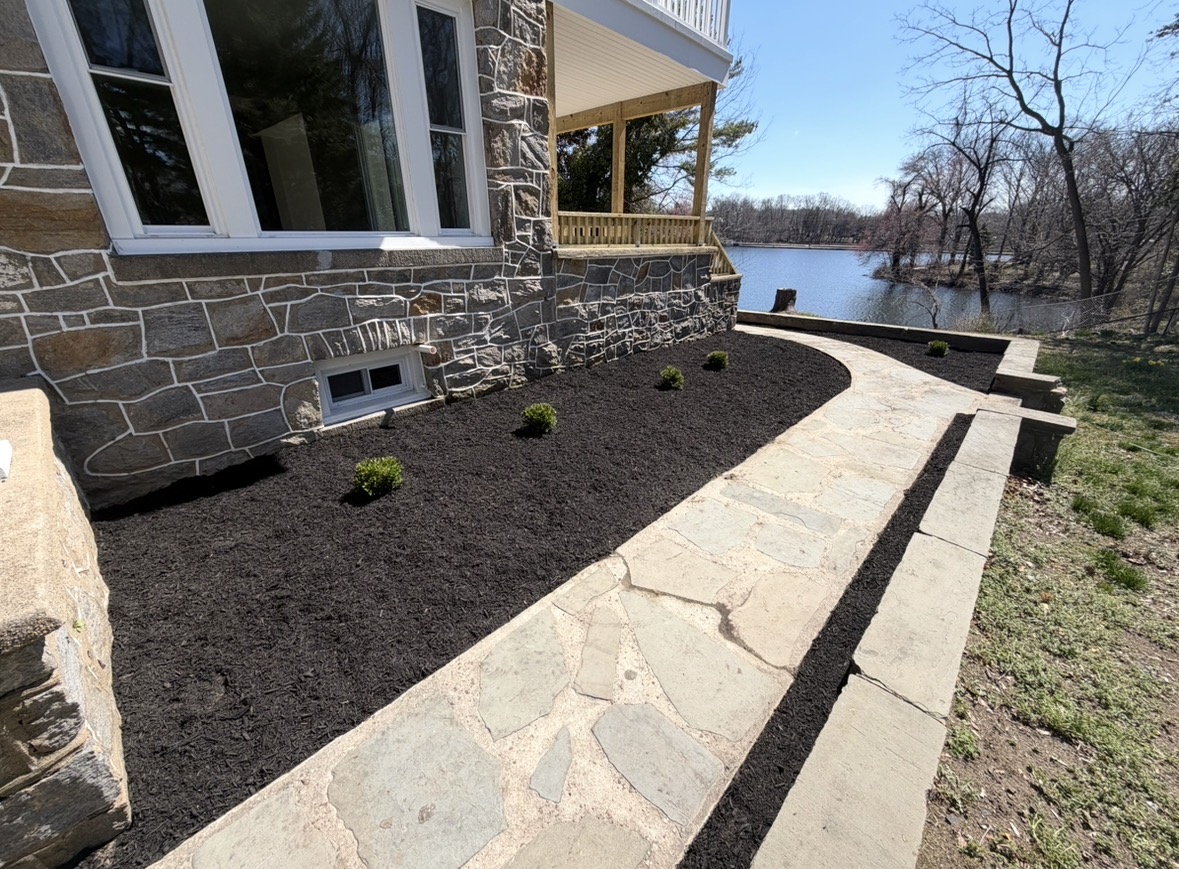 Flagstone walkway with stone wall and fresh mulch beds beside a stone home overlooking water